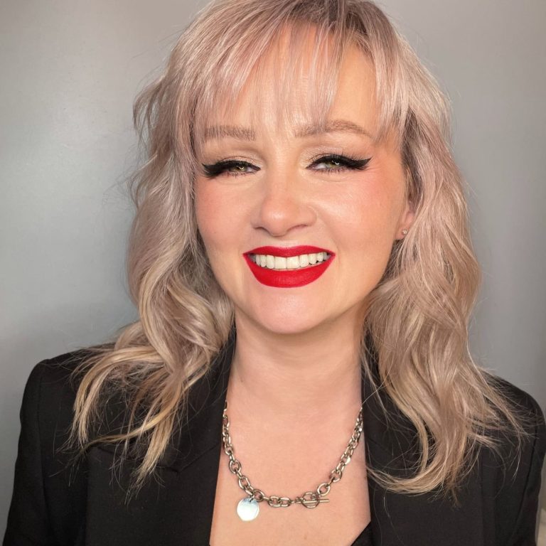 Lächelnde Frau mit blonden, lockigen Haaren und rotem Lippenstift, trägt schwarzen Blazer.