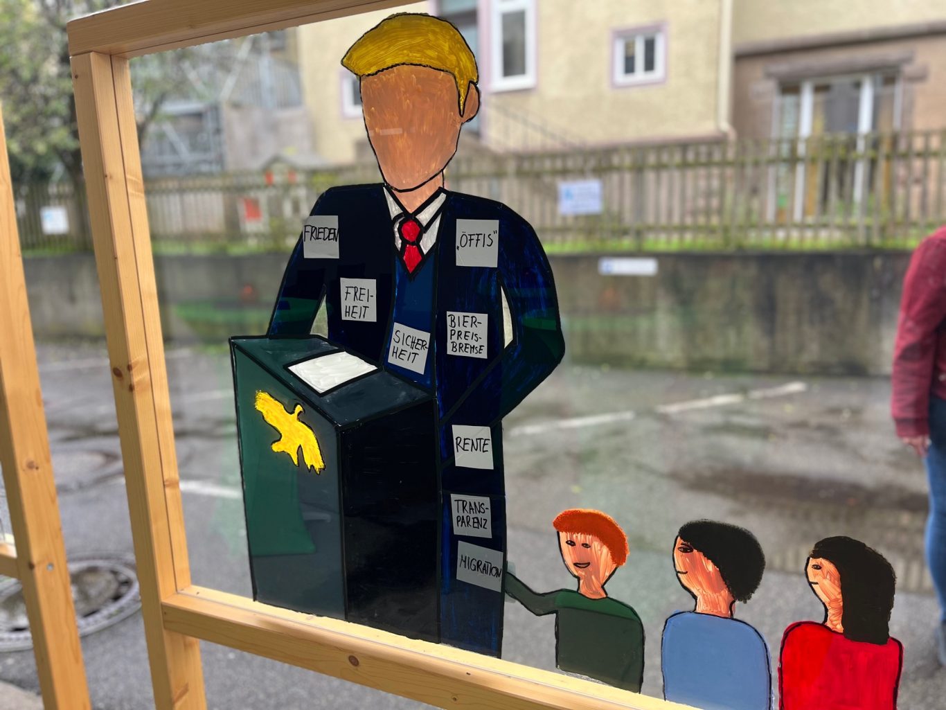 Stellwand mit einer Zeichnung von einem Politiker am Rednerpult, umgeben von drei Menschen, die ihm aufmerksam zuhören.