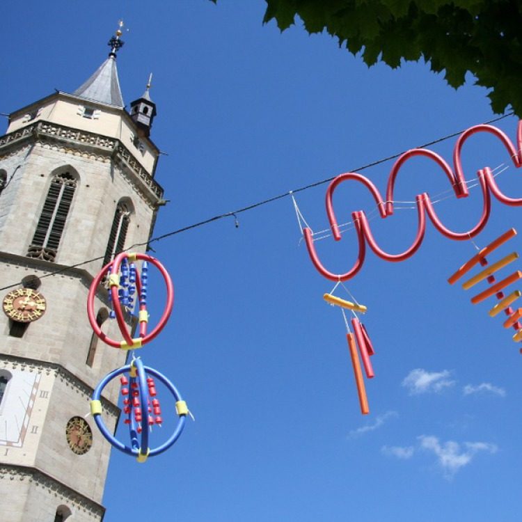 Farbige als Figuren gebogene Schwimmnudeln hängen an einem Draht neben einem Kirchturm vor klarem Himmel.