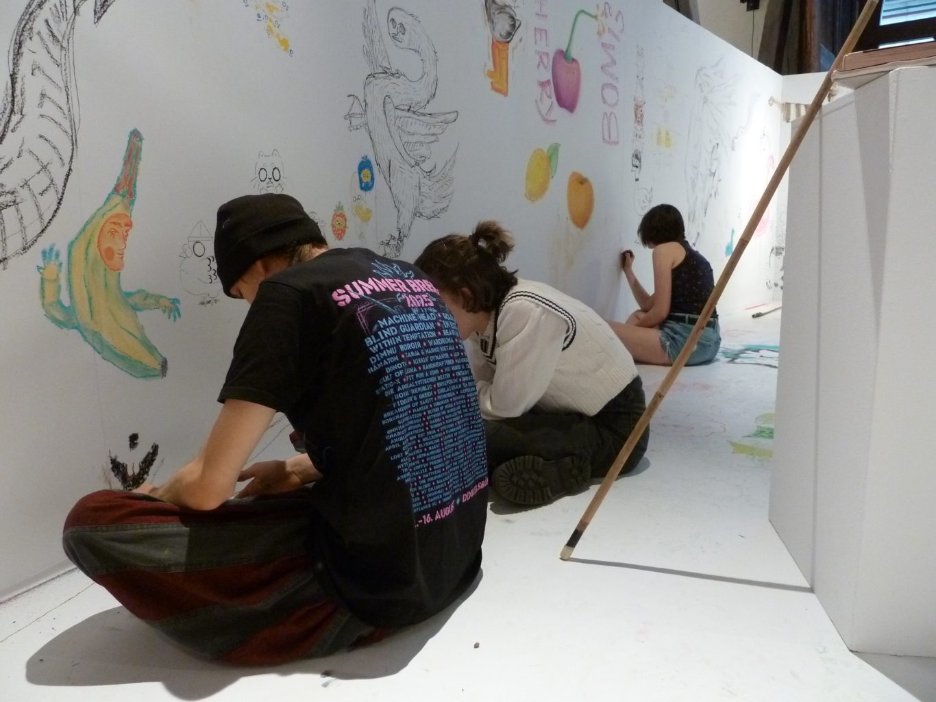 Drei Personen malen an einer langen, weißen Wand mit bunten Zeichnungen.