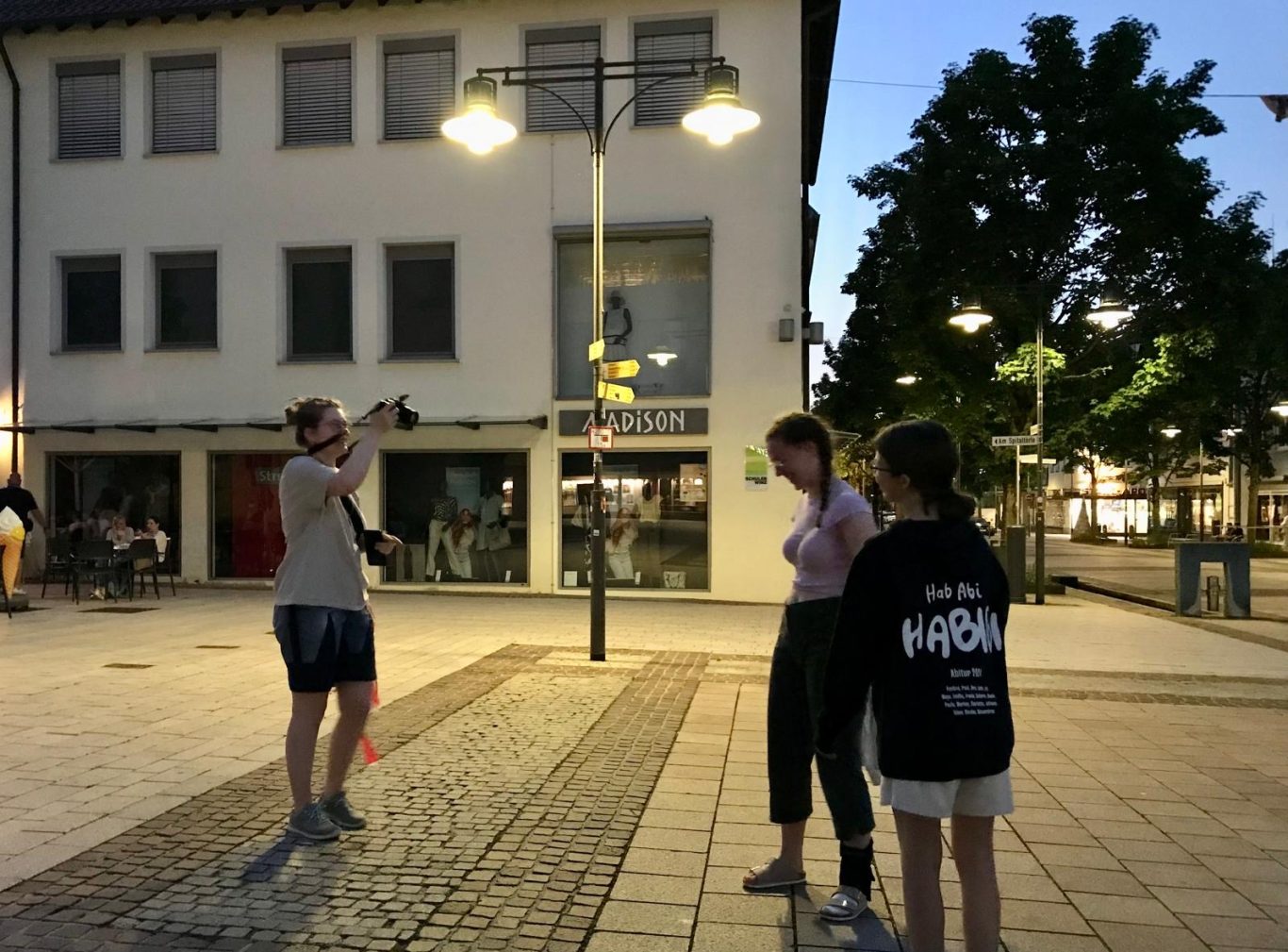 Drei Personen stehen auf einem Platz bei Nacht, umgeben von Laternen und Gebäuden.