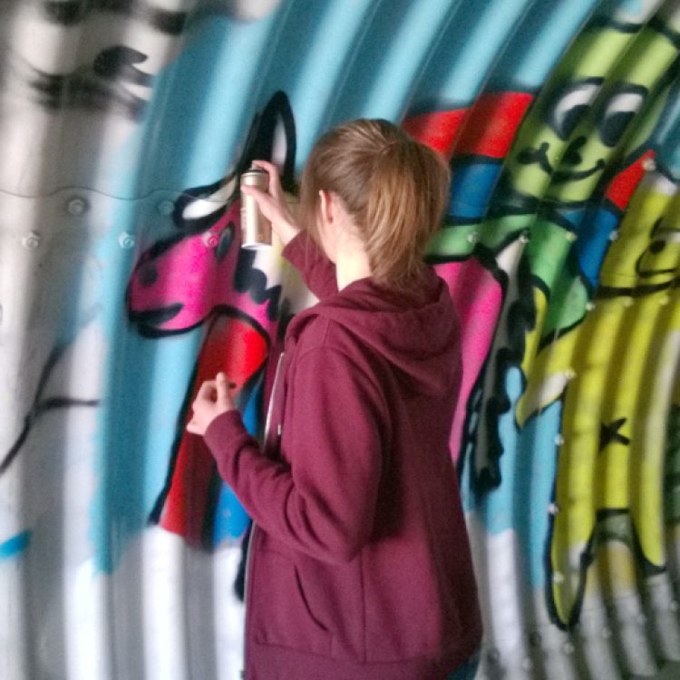 Junge Person bemalt eine Wand mit bunten Tiergrafiken in einem Tunnel.