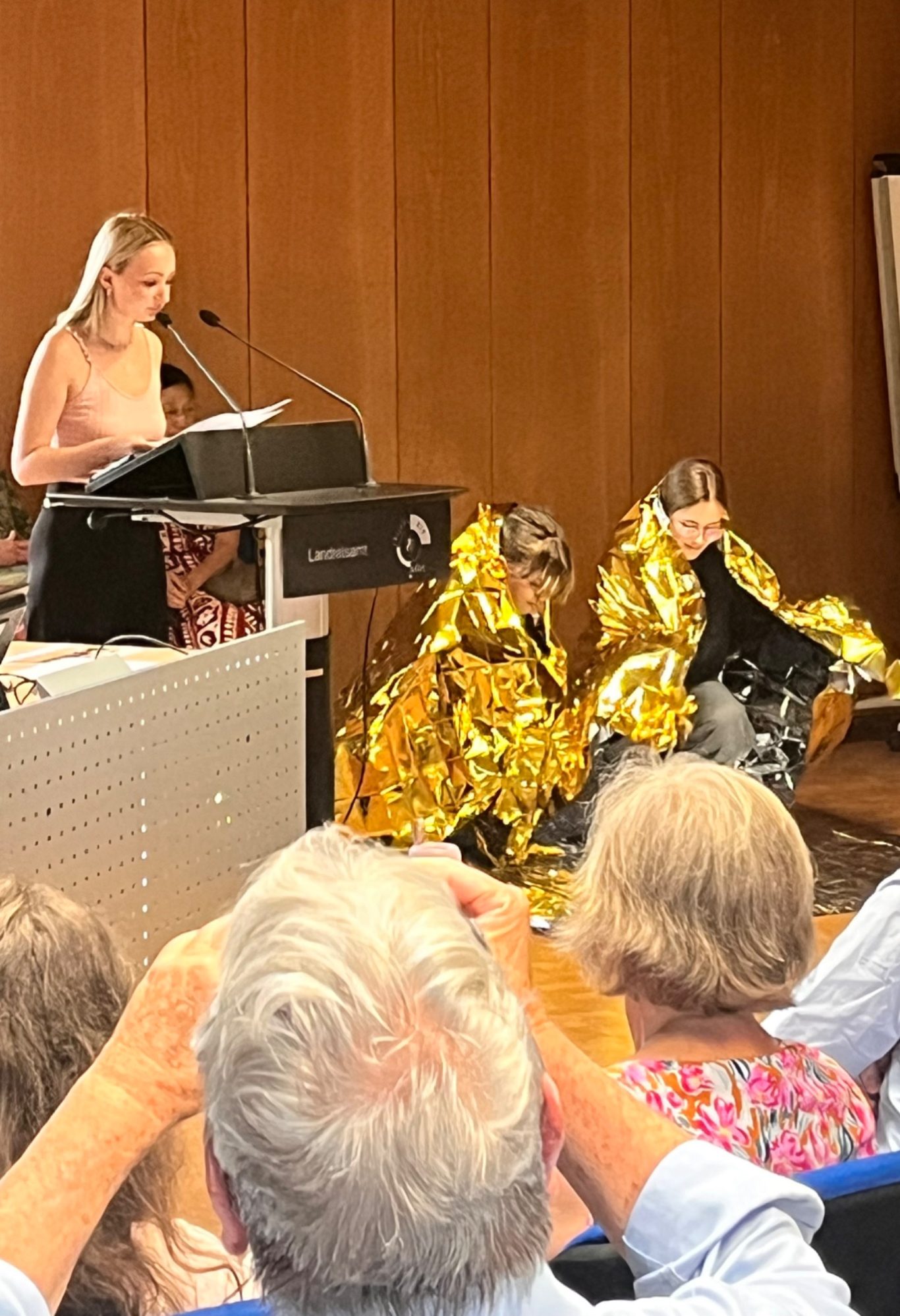 Eine Frau spricht am Mikrofon, während zwei Personen in goldenen Decken sitzen.