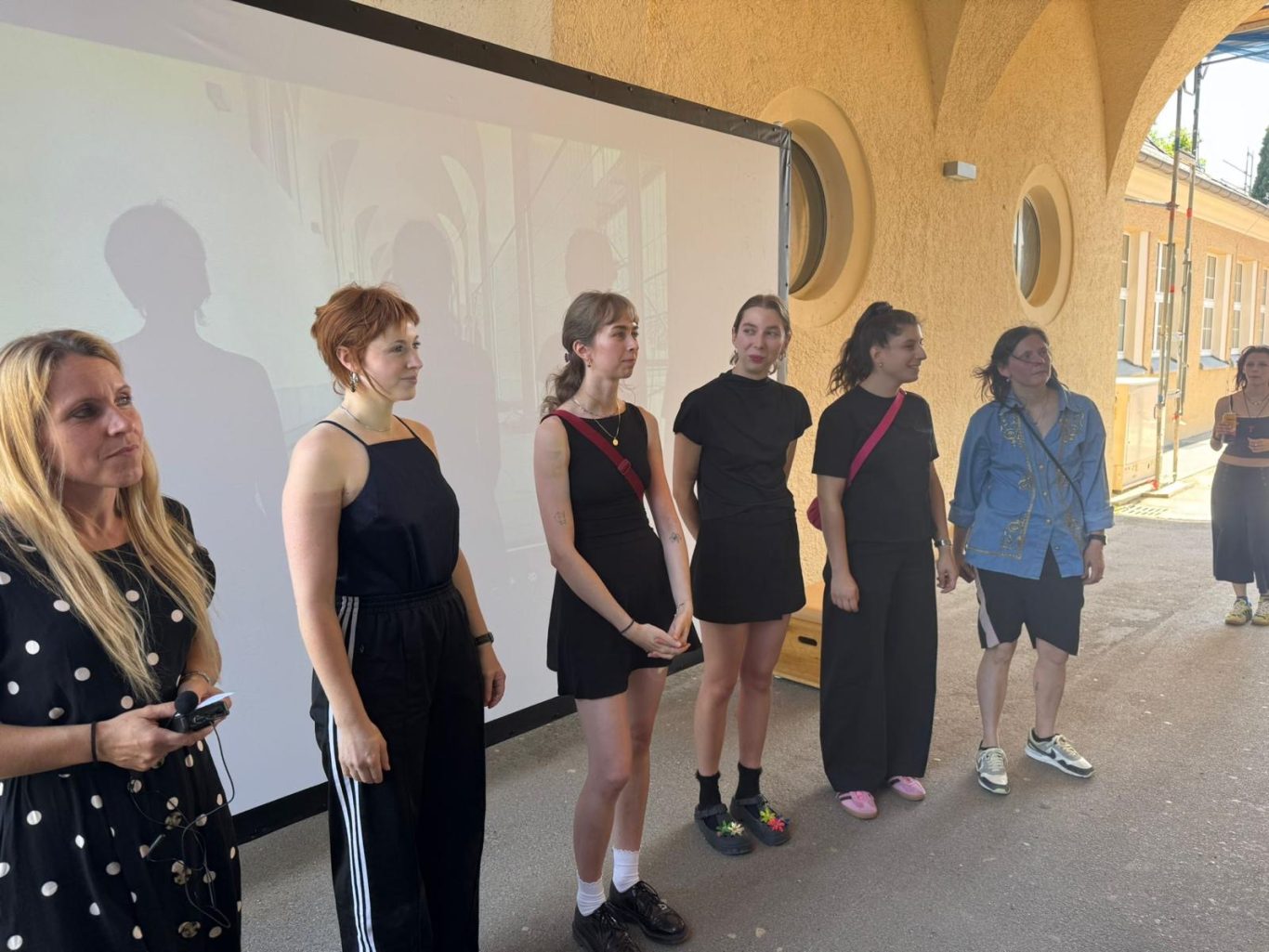 Sechs Personen stehen nebeneinander, einige sprechen, im Hintergrund ein Präsentationsbildschirm.