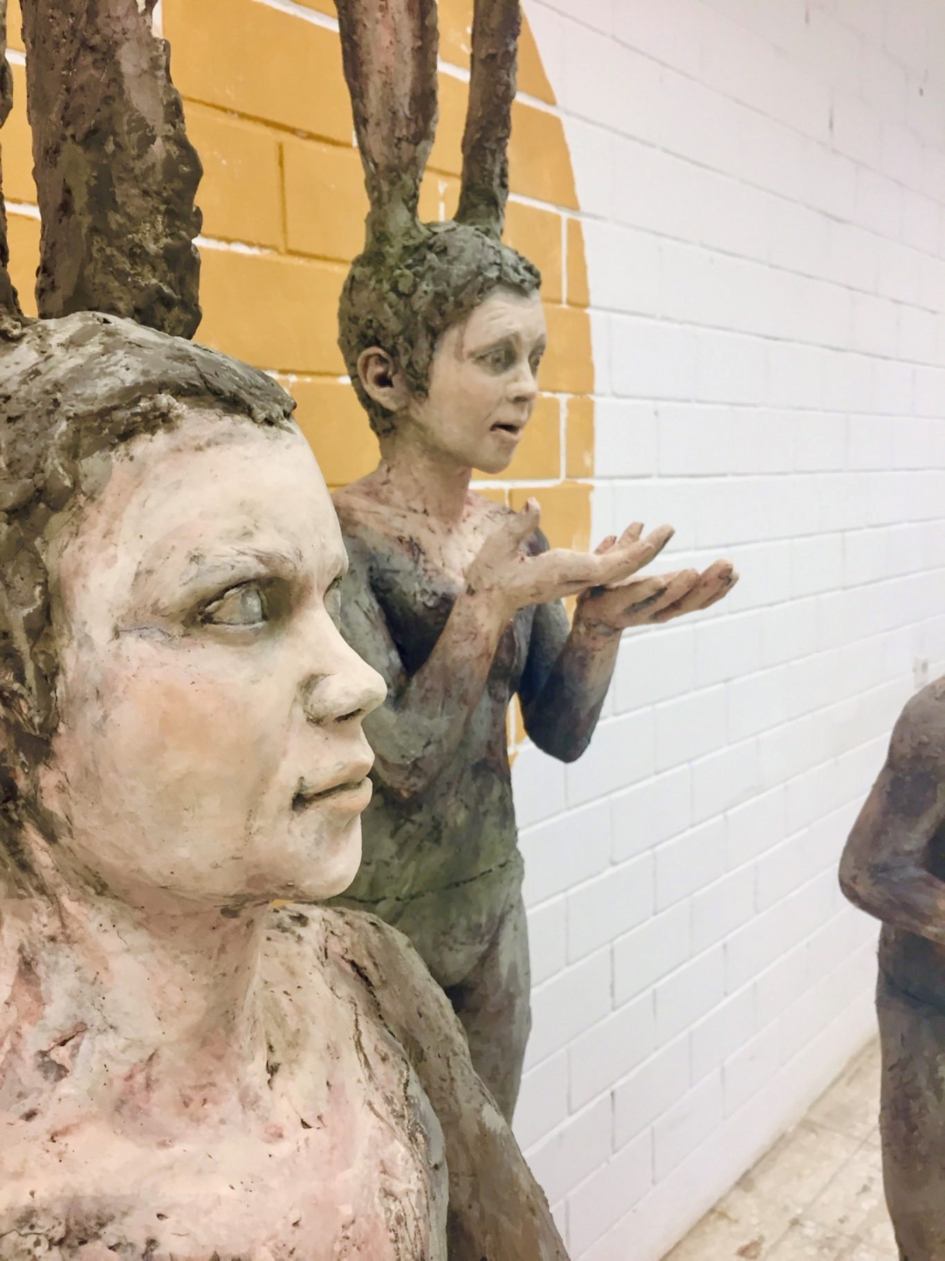 Skulpturen von menschlichen Figuren mit fantasievollen Merkmalen vor orangefarbener Wand.