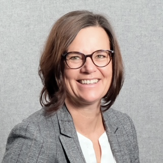 Frau mit Brille und grauem Blazer, lächelt freundlich vor neutralem Hintergrund.