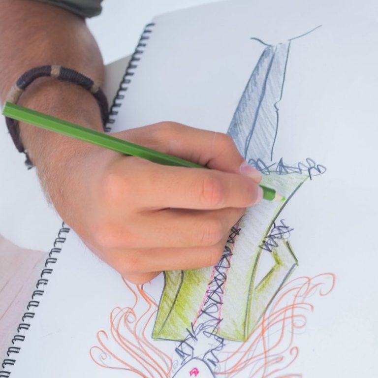 Eine Hand zeichnet mit einem grünen Bleistift eine bunte Modeillustration.