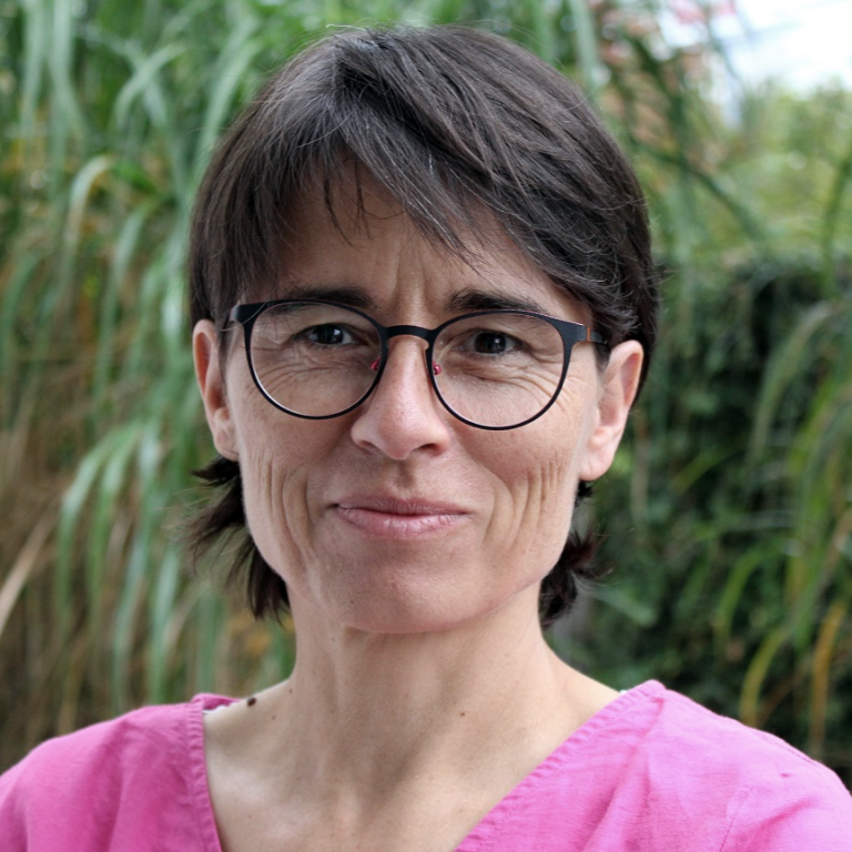 Frau mit Brille und kurzem Haar, lächelnd vor grüner Pflanzenkulisse.