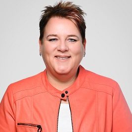 Frau mit Brille und kurzem Haar, lächelnd vor grüner Pflanzenkulisse.