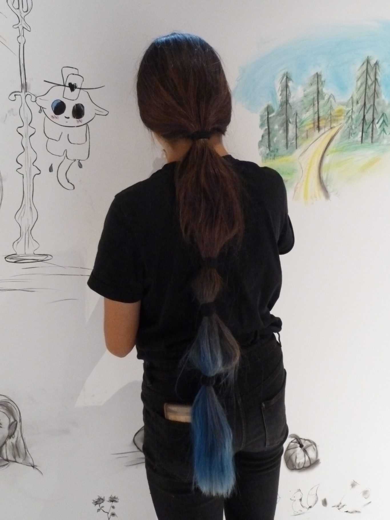 Person mit geflochtenem Haar in Schwarz und Blau, steht vor einer Wand mit Zeichnungen.