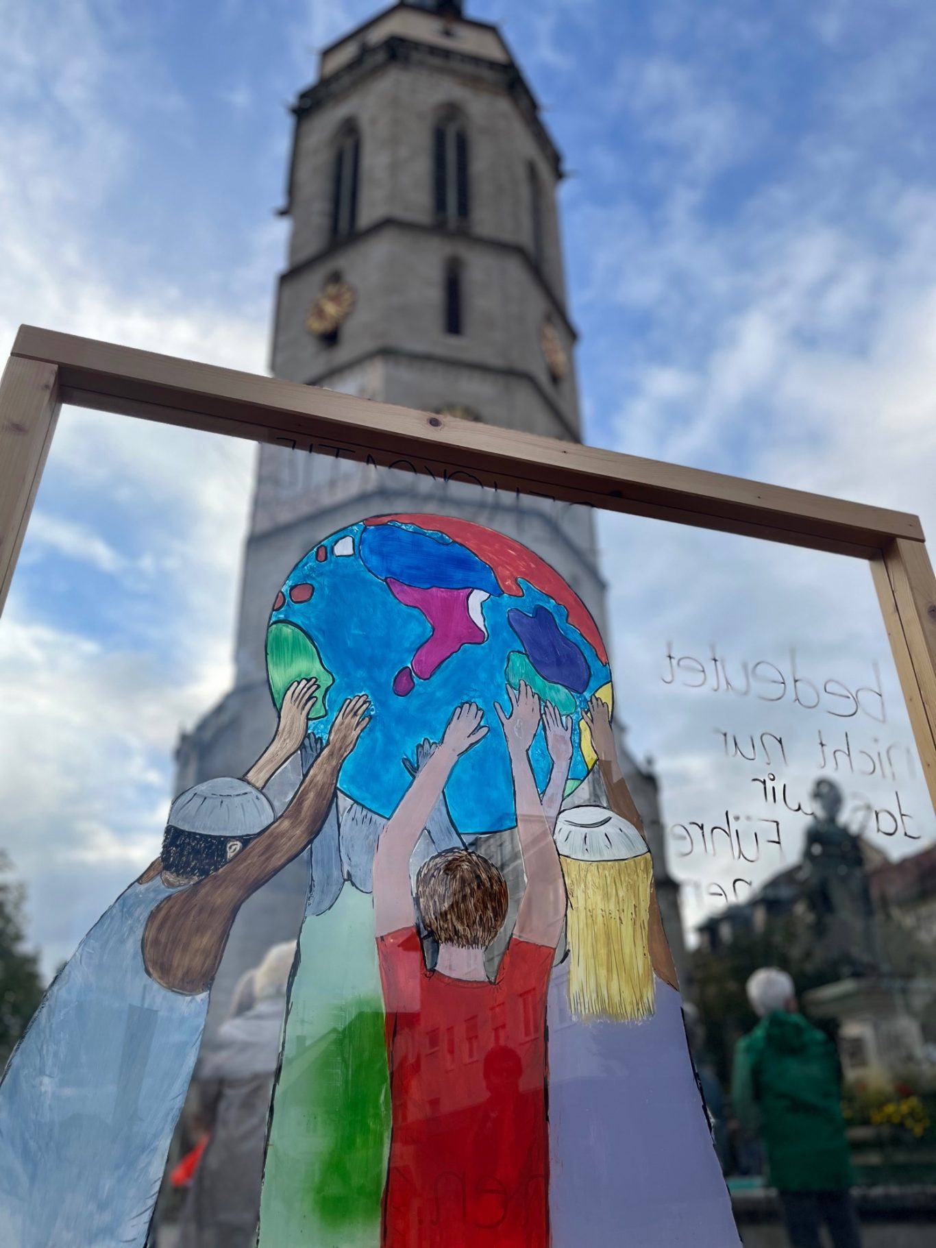 Bunte Stellwand mit einer Zeichnung mit Menschen, die eine Erde halten. Im Hintergrund ist ein Kirchturm zu sehen.