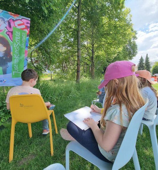 Kinder sitzen auf Stühlen im Freien und zeichnen, umgeben von Bäumen und einem Plakat.