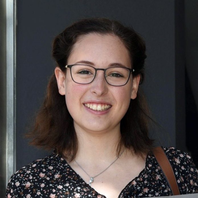 Junge Frau mit Brille und lockigem, braunem Haar, lächelnd vor dunklem Hintergrund.