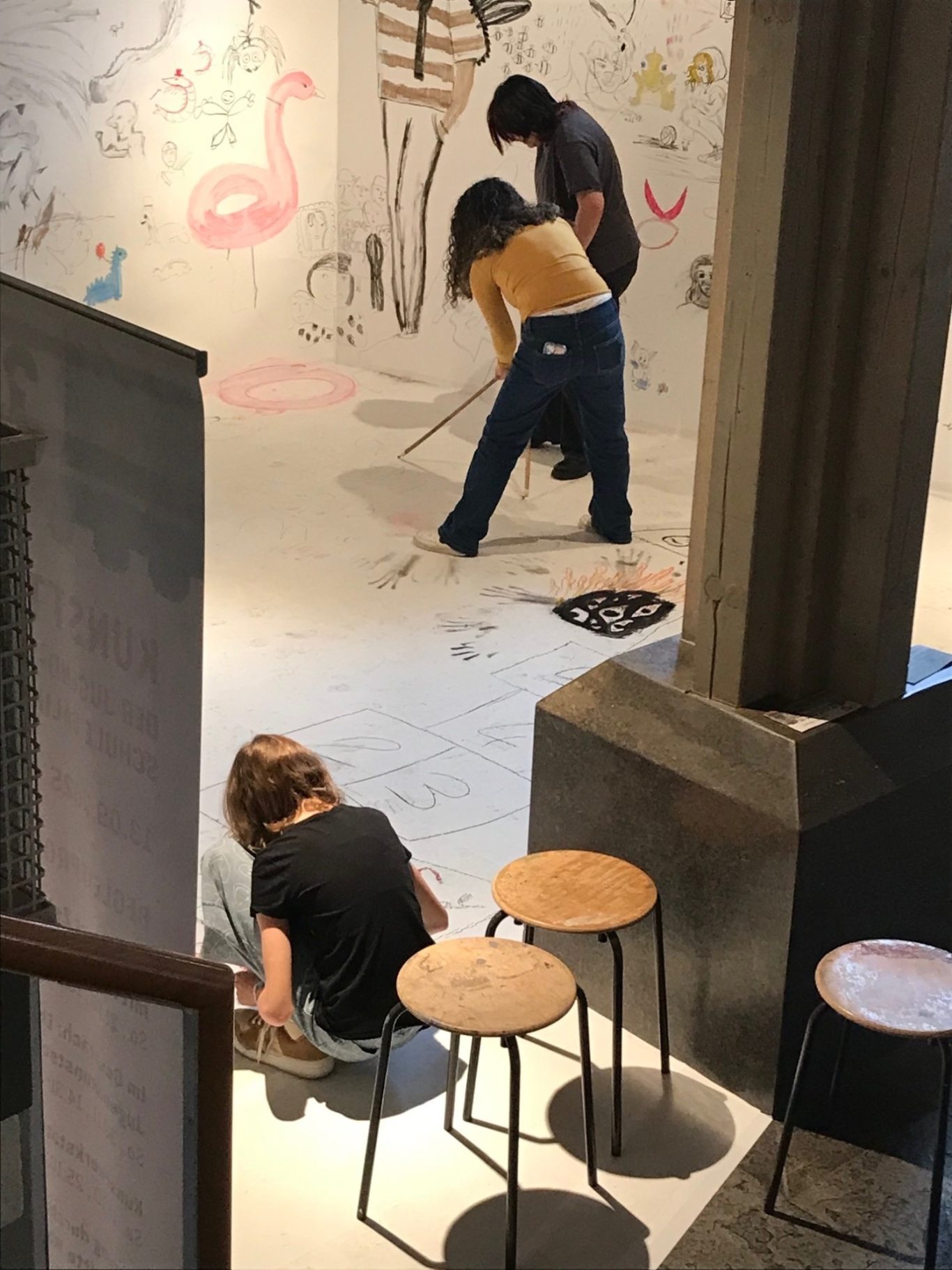 Drei Personen gestalten eine bemalte Wand in einem Innenraum. Holzstühle im Vordergrund.