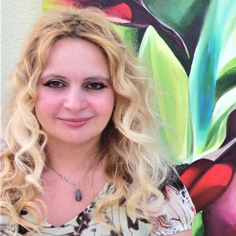 Eine lächelnde Frau mit lockigem, blonden Haar vor einer farbenfrohen Pflanzenmalerei.