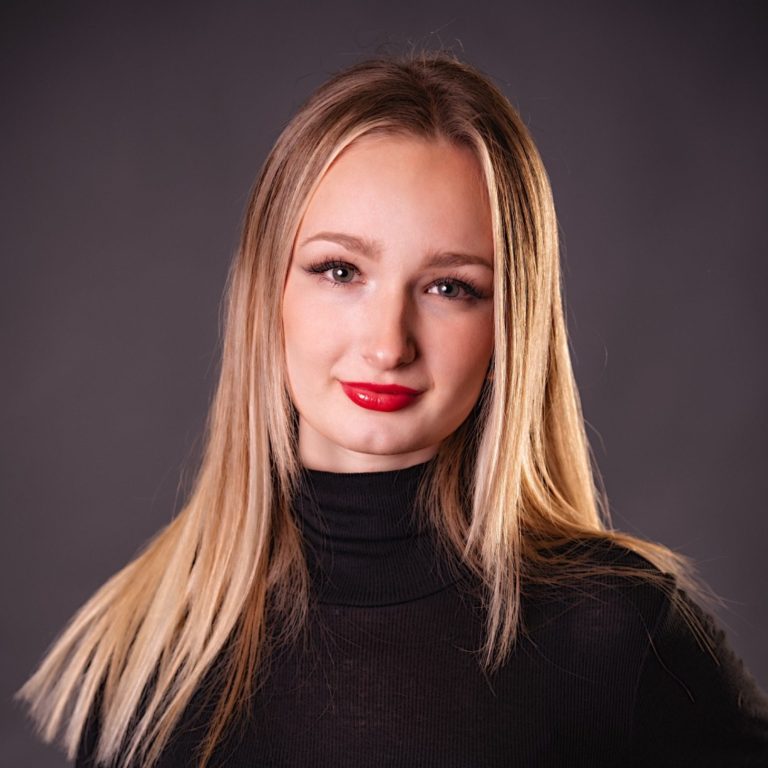 Frau mit Brille und kurzem Haar, lächelnd vor grüner Pflanzenkulisse.