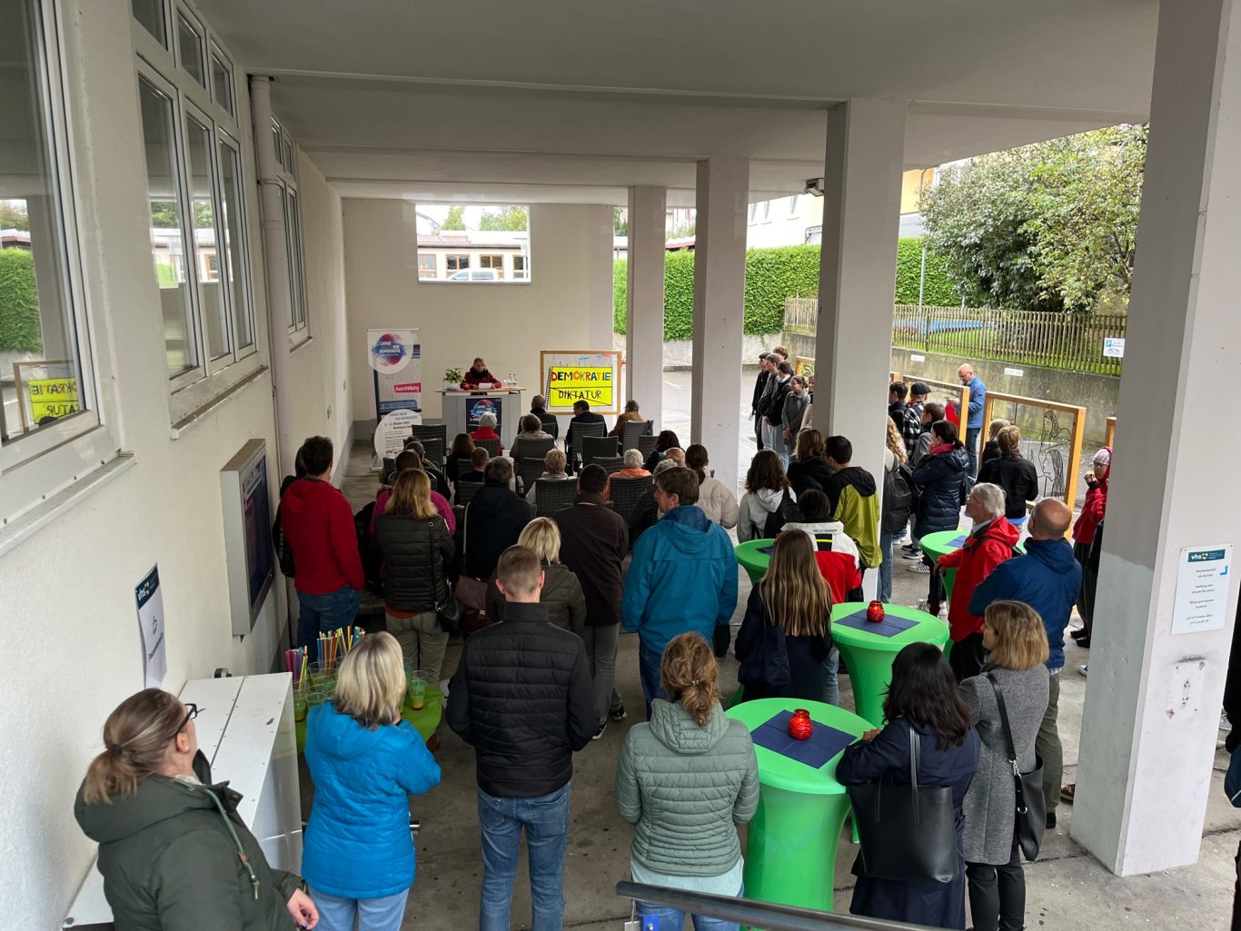 Menschenmenge versammelt sich im Freien, einige stehen an grünen Tischen.