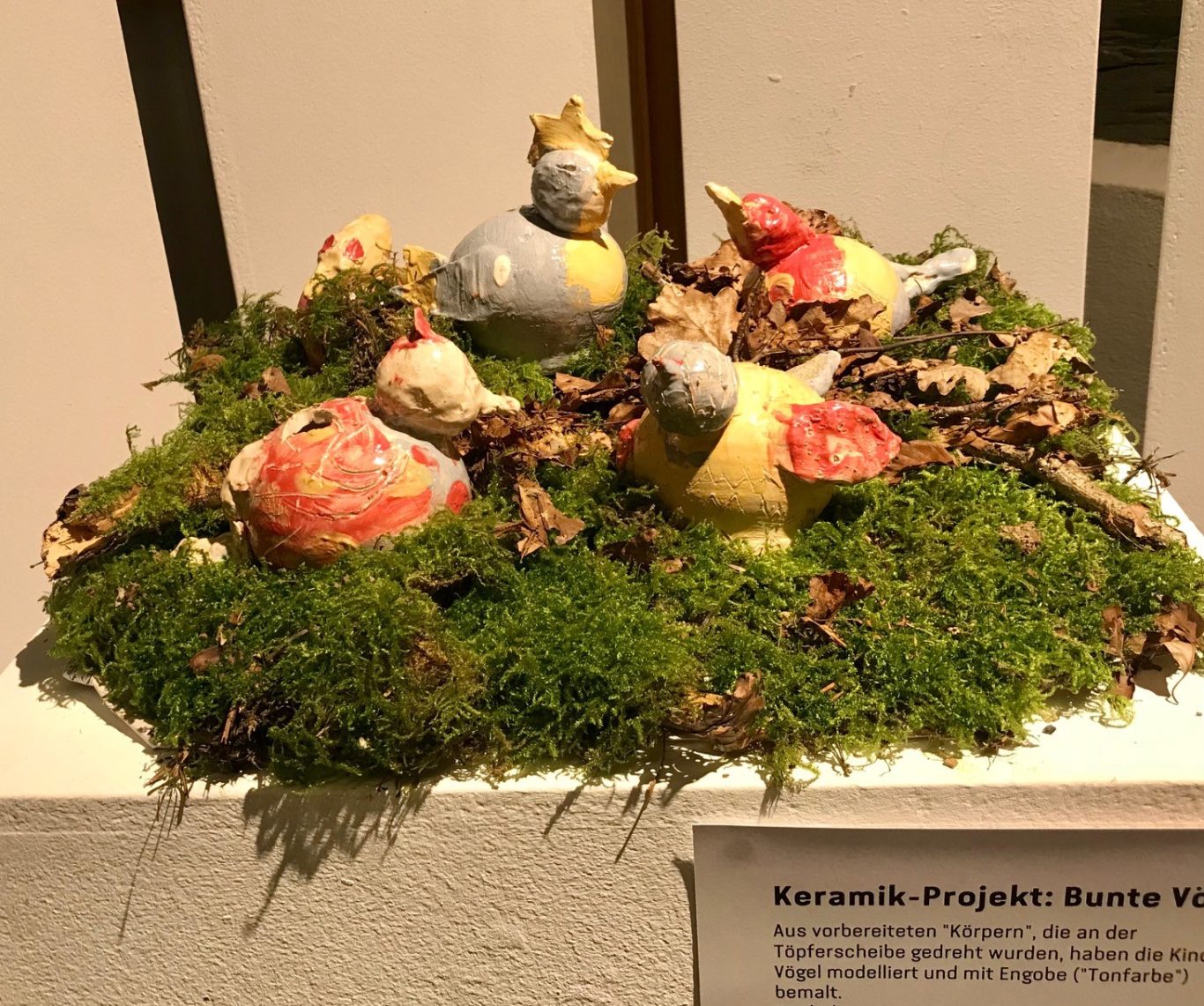Bunte Vögel aus Keramik auf Moos, umgeben von getrockneten Blättern.