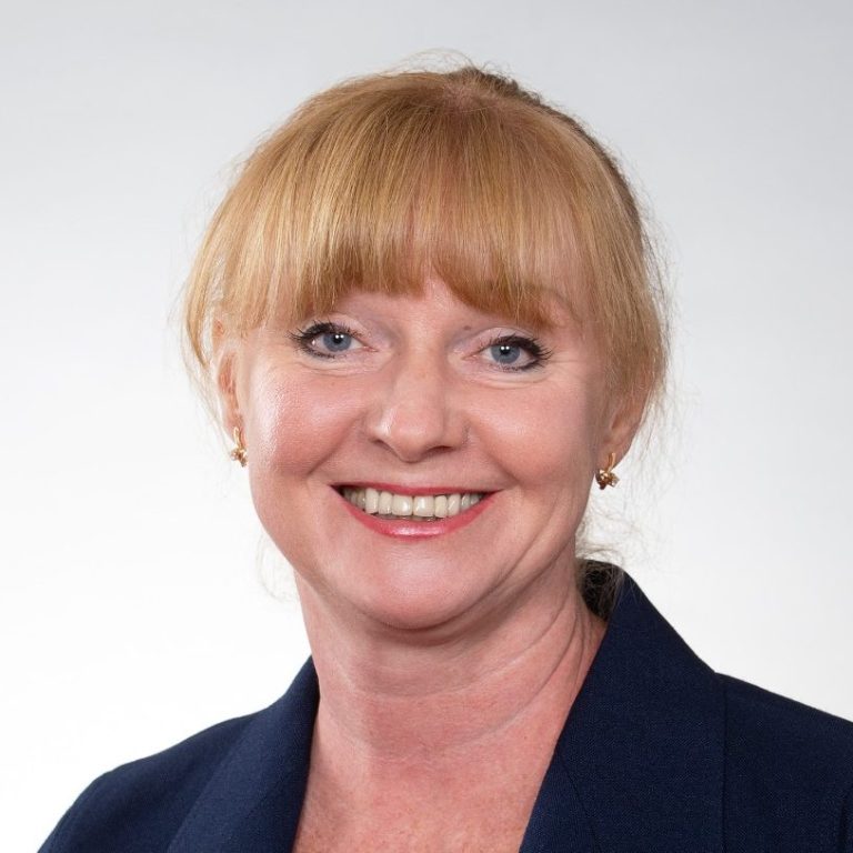 Lächelnde Frau mit blonden Haaren und blauer Jacke auf hellem Hintergrund.