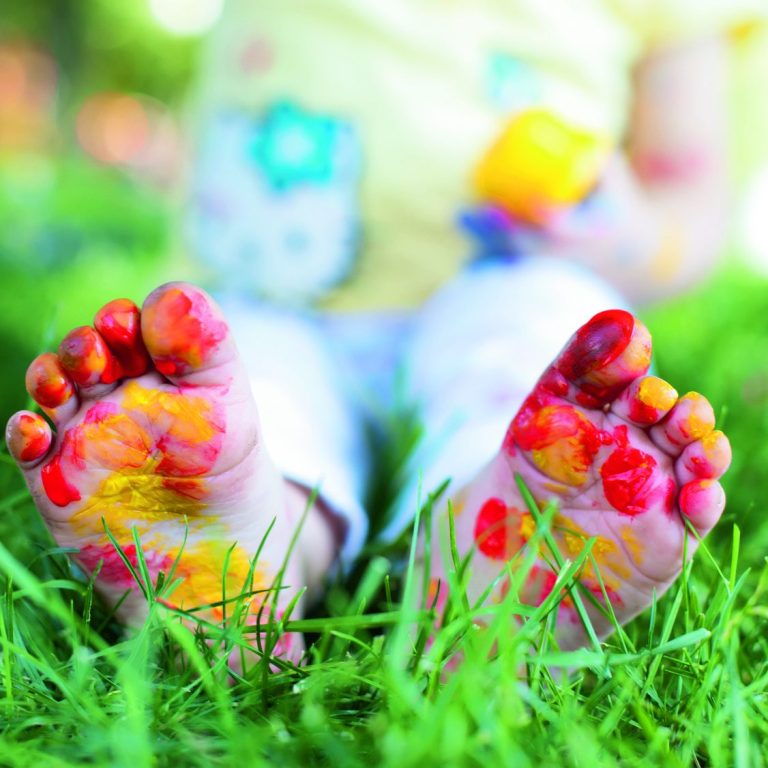 Bunte Füße eines Kindes mit Farbflecken, sitzend im Gras.