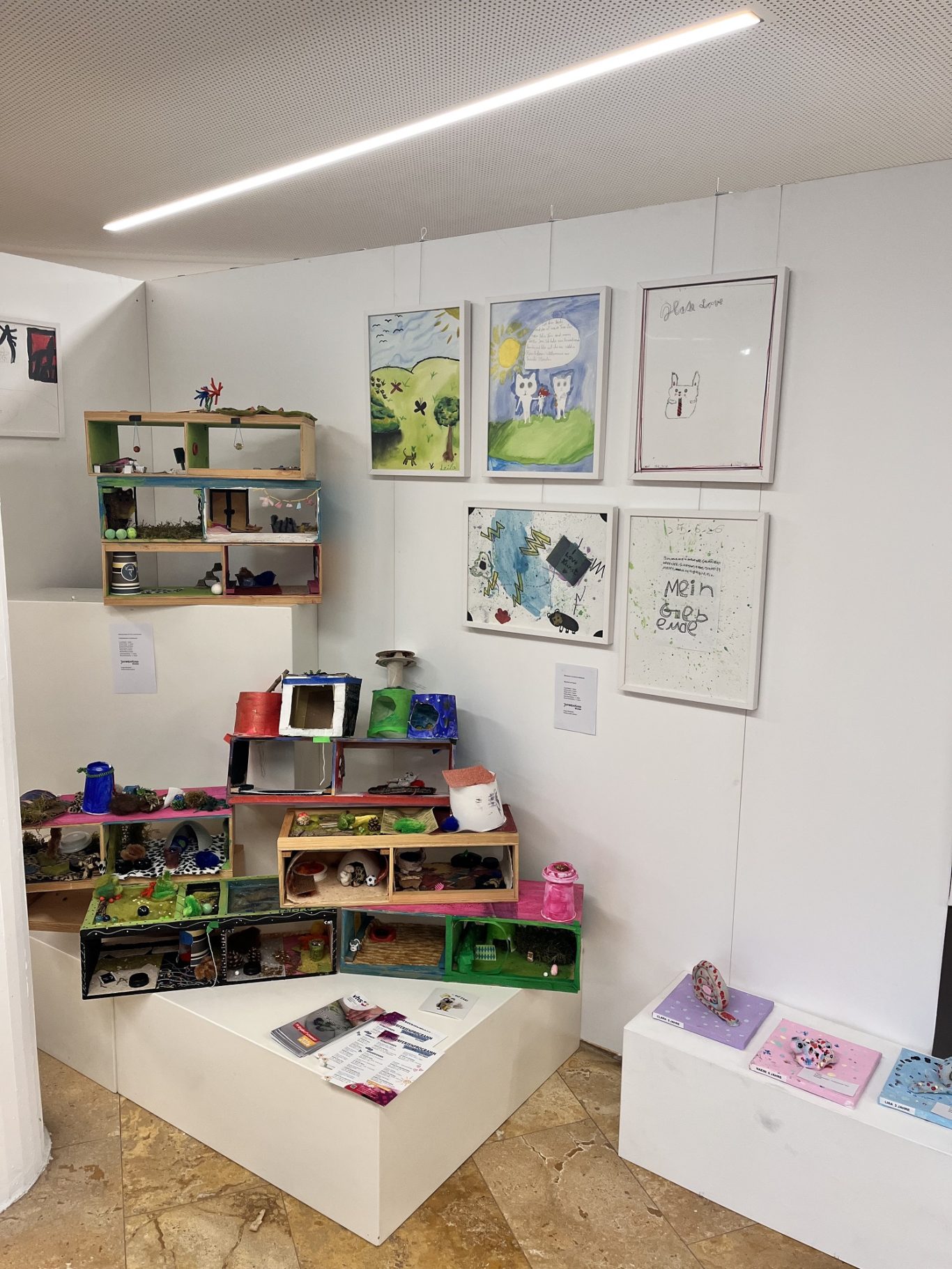 Ausstellung mit Regalen, Kunstwerken an der Wand und farbenfrohen Bastelmaterialien.