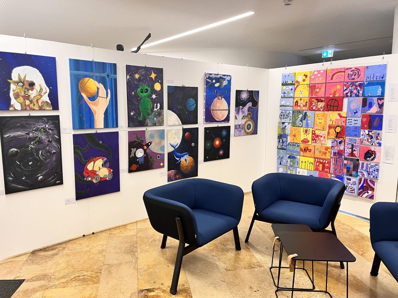 Farbenfrohe Wand mit verschiedenen abstrakten Kunstwerken und blauen Sesseln.